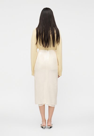 Suknja boje kreme, dužine do koljena, sjajne teksture i ravnog kroja, uparena s mekanom, svijetložutom majicom.