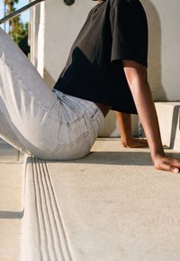 Personne portant un jean clair et une chemise noire assise sur des marches en béton extérieures avec bord texturé au soleil.