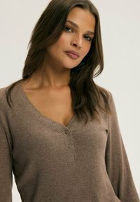 Brązowa, prążkowana bluzka z długim rękawem, z dekoltem w szpic i z detalami guziczków na klatce piersiowej. Posiada miękką teksturę i dopasowany krój.