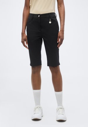 Personne portant un short noir mi-genou avec des poches latérales et un petit pendentif rond, associé à des chaussettes blanches et des baskets.