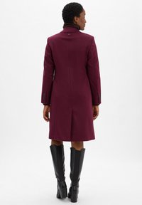 Vrouw in een knielange bordeauxrode jas en zwarte haklaarzen tot de knie, staande met haar rug naar de camera tegen een effen witte achtergrond.