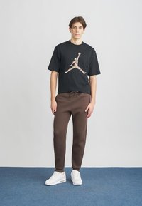 Junger Mann steht auf einem blauen Teppichboden vor einer schlichten hellen Wand und trägt ein schwarzes T-Shirt mit Grafik, braune Jogginghosen und weiße Turnschuhe.