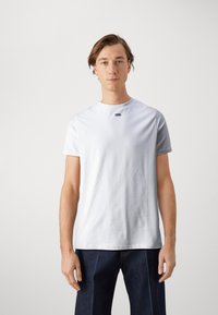 Camiseta de algodón blanca con cuello redondo, mangas cortas y un pequeño logo negro en el pecho. Llevada con pantalones de mezclilla azul oscuro.