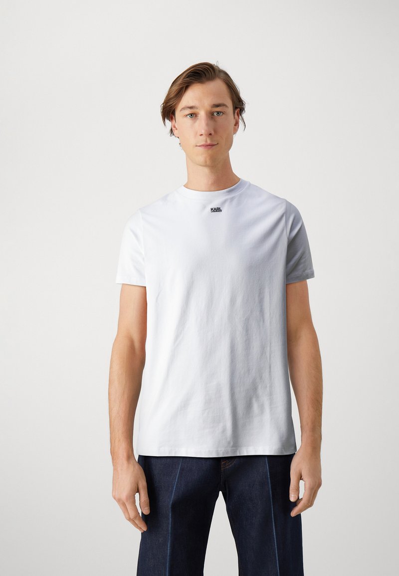 Camiseta de algodón blanca con cuello redondo, mangas cortas y un pequeño logo negro en el pecho. Llevada con pantalones de mezclilla azul oscuro.