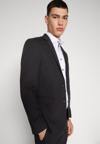 Black suit jacket featuring a notched lapel, two buttons, and side pockets. Worn over a white shirt with black buttons and a blue accent on the collar.