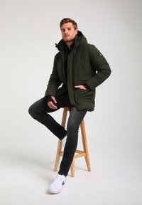 Giacca a vento verde oliva con cappuccio, bottoni frontali e tasche laterali. Abbinata a jeans scuri e sneakers bianche su uno sgabello di legno.
