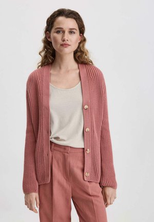 Roze geribbelde cardigan met houten knopen, gedragen over een lichtbeige top. Het model combineert het met bijpassende roze broeken. Gladde textuur.