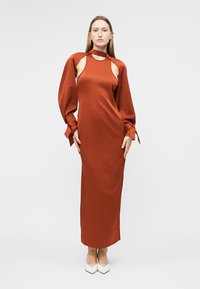 Vrouw staand, gekleed in een lang roestoranje jurk met openingen op de schouders en lange opgeblazen mouwen die aan de polsen zijn gestrikt, gecombineerd met witte puntige schoenen.