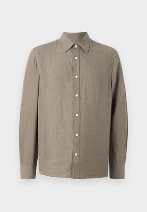 Mand iført olivengrøn skjorte med knapper og beige vidde bukser, holder en brun lædertaske og står mod en ensfarvet lys baggrund.