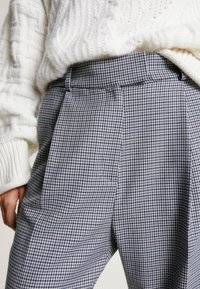 Grijze en witte houndstooth-patronen broeken met een hoge taille, geplooid ontwerp en zijzakken, gecombineerd met een chunky witte gebreide sweater.