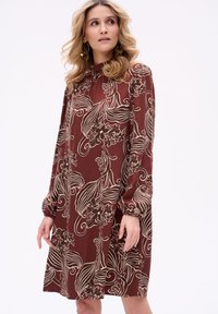 Czekoladowa długa sukienka w kwiaty z długimi rękawami, wysokim kołnierzem, bufkami oraz misternymi białymi wzorami kwiatowymi na jedwabnym materiale.