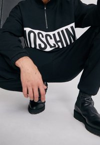 Černá mikina na zip s výrazným bílým logem "MOSCHINO" na hrudi, doplněná o fitted černé kalhoty a černé kotníkové boty.
