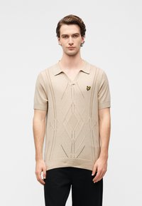 Μπεζ πουλόβερ με κοντά μανίκια, με V-λαιμόκοψη, υφασμένο γεωμετρικό μοτίβο και μικρό κίτρινο λογότυπο πεταλούδας στο στήθος.