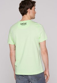 Camiseta de algodón verde claro con cuello redondo y mangas cortas. Presenta un logo impreso en la parte posterior cerca del cuello.