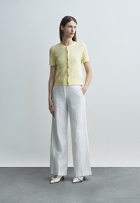 Femeie stând în picioare, purtând un cardigan galben deschis cu mânecă scurtă și nasturi, pantaloni albi cu picioare largi și pantofi albi cu vârf ascuțit.