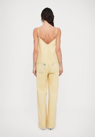 Γυναίκα με σκούρα μαλλιά, φοράει αμάνικη ολόσωμη φόρμα ανοιχτού κίτρινου χρώματος, άνετη και με χαμηλή πλάτη και δύο πίσω τσέπες, στέκεται μπροστά σε λευκό φόντο.