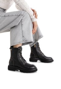 Botines de cuero negro con diseño de cordones, ojales dorados, cremallera lateral, tachuelas metálicas en los lados y suelas de goma gruesa.