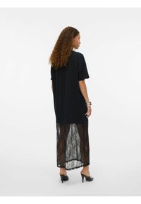 Femme aux cheveux bouclés portant un t-shirt noir surdimensionné et une jupe en dentelle noire transparente, se tenant de côté avec des chaussures à talons sur fond blanc.