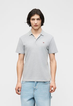 Νεαρός άνδρας με μεσαίου μήκους σκούρα μαλλιά, φοράει ανοιχτό γκρι κοντομάνικο πόλο πουκάμισο και ανοιχτό μπλε τζιν, μπροστά σε απλό φόντο.