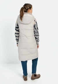 Lang, lys beige puffer vest med hætte, der har horisontale quiltede sektioner og en blød tekstur, båret over en mønstret sweater og blå jeans.