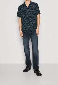 Chemise à manches courtes à boutons, noire avec motif abstrait turquoise ; associée à un jean foncé et des chaussures noires. Texture lisse, coupe décontractée.