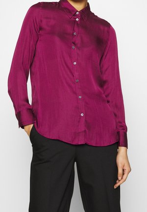 Personne portant une chemise satinée fuchsia à manches longues boutonnée, rentrée dans un pantalon noir, avec une main dans la poche, sur un fond uni.