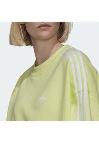 Žlutý svetr s bílými třemi pruhy na ramenou. Má kulatý výstřih a vyšívané logo na přední straně. Složení z bavlny.