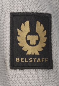 Černá tkaninová nášivka se zlatým emblémem s křídly a kruhovým designem. Text "BELSTAFF" ve zlaté barvě dole, umístěný na šedém pozadí.