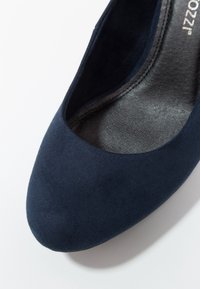 Gros plan sur une chaussure plate en daim bleu marine avec un bout rond et une semelle intérieure noire rembourrée sur fond blanc.