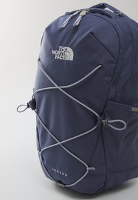 Marineblå ryggsekk med teksturert materiale, elastisk snor-design, sidemesh-lomme og logo-trykk; har grå detaljer og justerbare stropper.