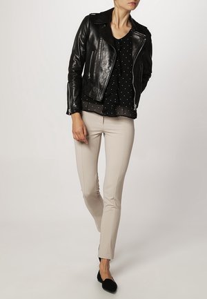 Giacca da biker in pelle nera, design aderente, cerniere argentate, abbinata a una blusa nera con pois e pantaloni beige su misura. Scarpe basse nere.