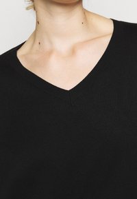 Gros plan d'une personne portant un pull noir à encolure en V, montrant la ligne du cou, les clavicules et la partie supérieure de la poitrine.