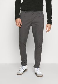 Man som bär mörkgrå upprullade byxor, vita sneakers med svarta ränder och en svart långärmad ribbad tröja, står mot en vit bakgrund.