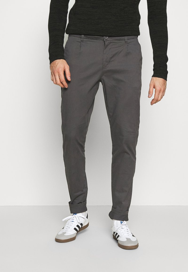 Man som bär mörkgrå upprullade byxor, vita sneakers med svarta ränder och en svart långärmad ribbad tröja, står mot en vit bakgrund.
