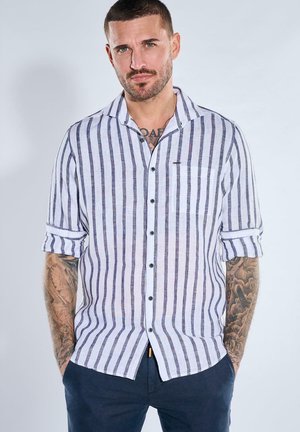 Man with short hair and tattoos on arms wearing a white button-up shirt with vertical black stripes and dark pants, hands in pockets.