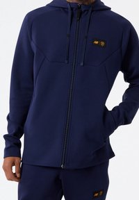 Felpa con zip navy realizzata in materiale morbido, con cappuccio, tasche frontali e toppe con logo a contrasto sul petto e sulle maniche.