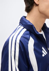 Κοντινό πλάνο ενός ατόμου που φορά αθλητικό μπουφάν με φερμουάρ σε μπλε χρώμα, με λευκές ρίγες στους ώμους και λευκό λογότυπο στο στήθος.