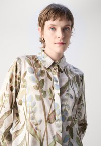 Camicia leggera con bottoni realizzata in tessuto con un motivo floreale nei colori verde, blu e beige. Collo classico e maniche lunghe.