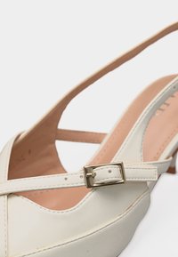 Chaussure slingback en cuir blanc pour femme avec boucle dorée sur la bride et petit talon, vue en gros plan sur fond blanc.
