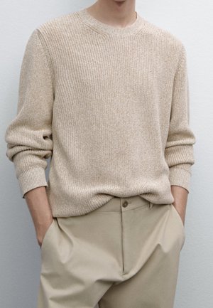 Personne portant un pull en laine beige avec les manches retroussées et un pantalon beige clair, debout les mains dans les poches devant un mur uni.