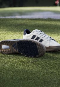 Λευκά και μαύρα αθλητικά παπούτσια με υφασμένα λαστιχένια πέλματα, που βρίσκονται πάνω σε πράσινο χορτάρι κοντά σε σημαία οπής γκολφ.