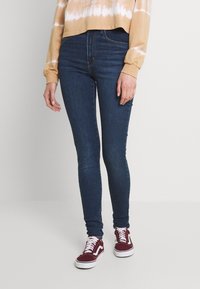 Jeans ajustados de color azul oscuro con una textura suave. Llevados con una camiseta de manga larga recortada con un diseño de tie-dye y zapatillas de ante burdeos.
