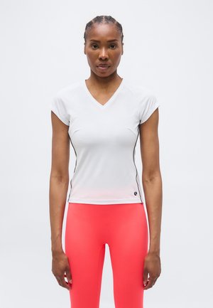 Kõrge naine palmikutega, seljas valge V-kaelusega sobiv T-särk ja erksad korallpunased retuusid, seismas lihtsa hele tausta ees.