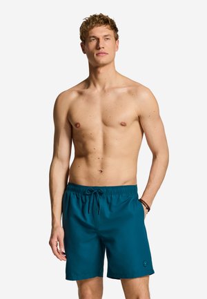 Jonge man staat zonder shirt, draagt turquoise zwemshort met trekkoord en heeft handen in de zakken tegen een effen achtergrond.