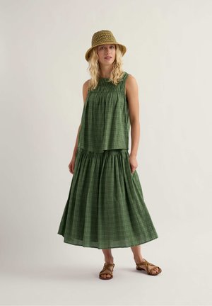 Femeie purtând un top verde fără mâneci și o fustă lungă asortată, pălărie maro țesută și sandale maro cu barete, stând în fața unui fundal simplu.