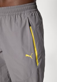 Shorts de sport gris avec des rayures latérales jaunes et le logo Puma, taille élastique, portés par une personne sur un fond uni.