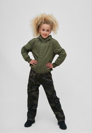 Zeleno-olivová mikina na zips s kapucňou, v kombinácii s čiernymi maskáčovými cargo nohavicami s vreckami. Model stojí sebavedomo s rukami na bokoch.