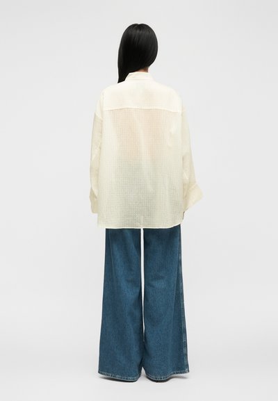 Femme aux longs cheveux noirs portant une chemise ample couleur crème et un jean bleu à jambes larges, se tenant face à un fond uni gris clair.