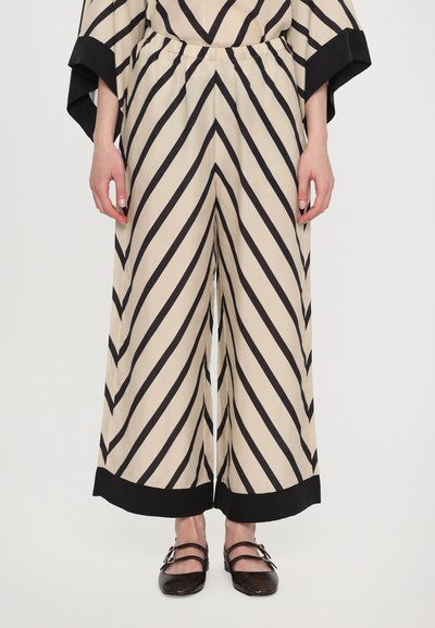 Pantalones de pierna ancha beige con rayas diagonales negras, cintura elástica y detalles en el dobladillo negros. Combinados con zapatos oscuros y texturizados.