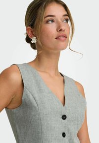 Jeune femme aux cheveux châtain clair attachés en chignon, portant de grandes boucles d'oreilles argentées florales et un haut gris sans manches à boutons avec un col en V.